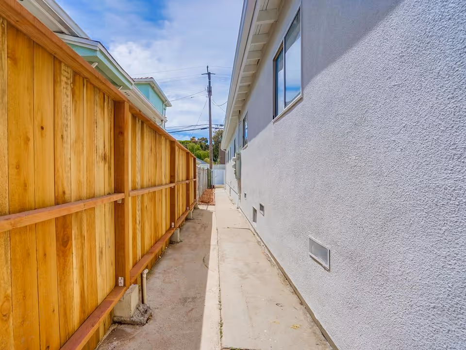 Narrow outdoor walkway between a white stucco building wall with windows and a wooden fence under a partly cloudy sky.