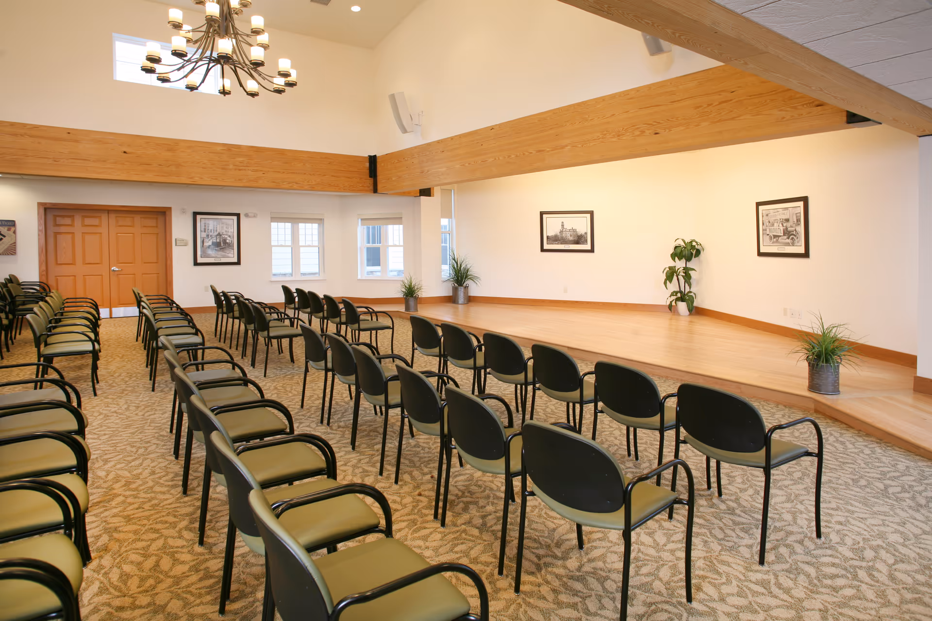A spacious room with rows of green and black chairs arranged facing a wooden stage. The room has beige patterned carpet, white walls with framed pictures, large wooden beams, and a chandelier hanging from the ceiling. There are also potted plants on and near the stage.