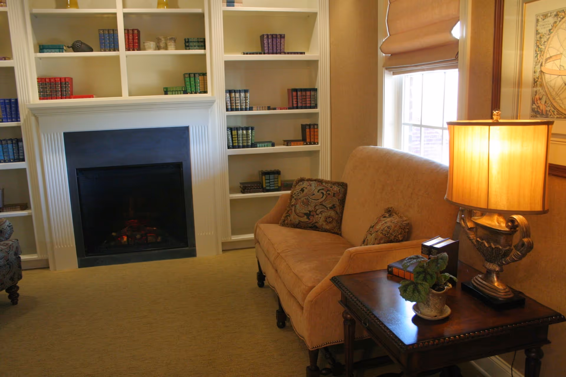 Cozy sitting area with a sofa and side table with a lamp beside a window, facing a fireplace flanked by built-in bookshelves.