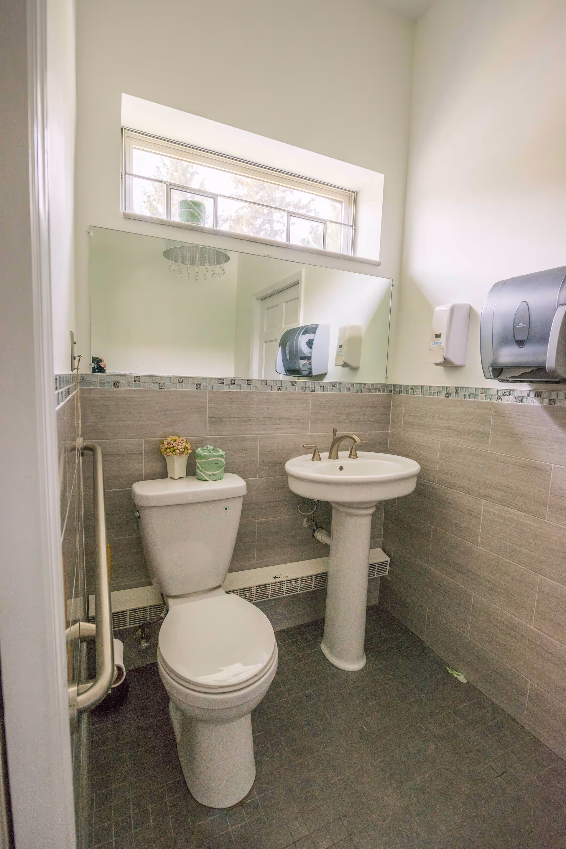 A clean and well-lit bathroom with a white toilet and pedestal sink. The walls are tiled halfway up with light gray tiles and a decorative border. Above the sink is a large mirror and a small window letting in natural light. There are wall-mounted soap and paper towel dispensers, and a grab bar next to the toilet for accessibility.