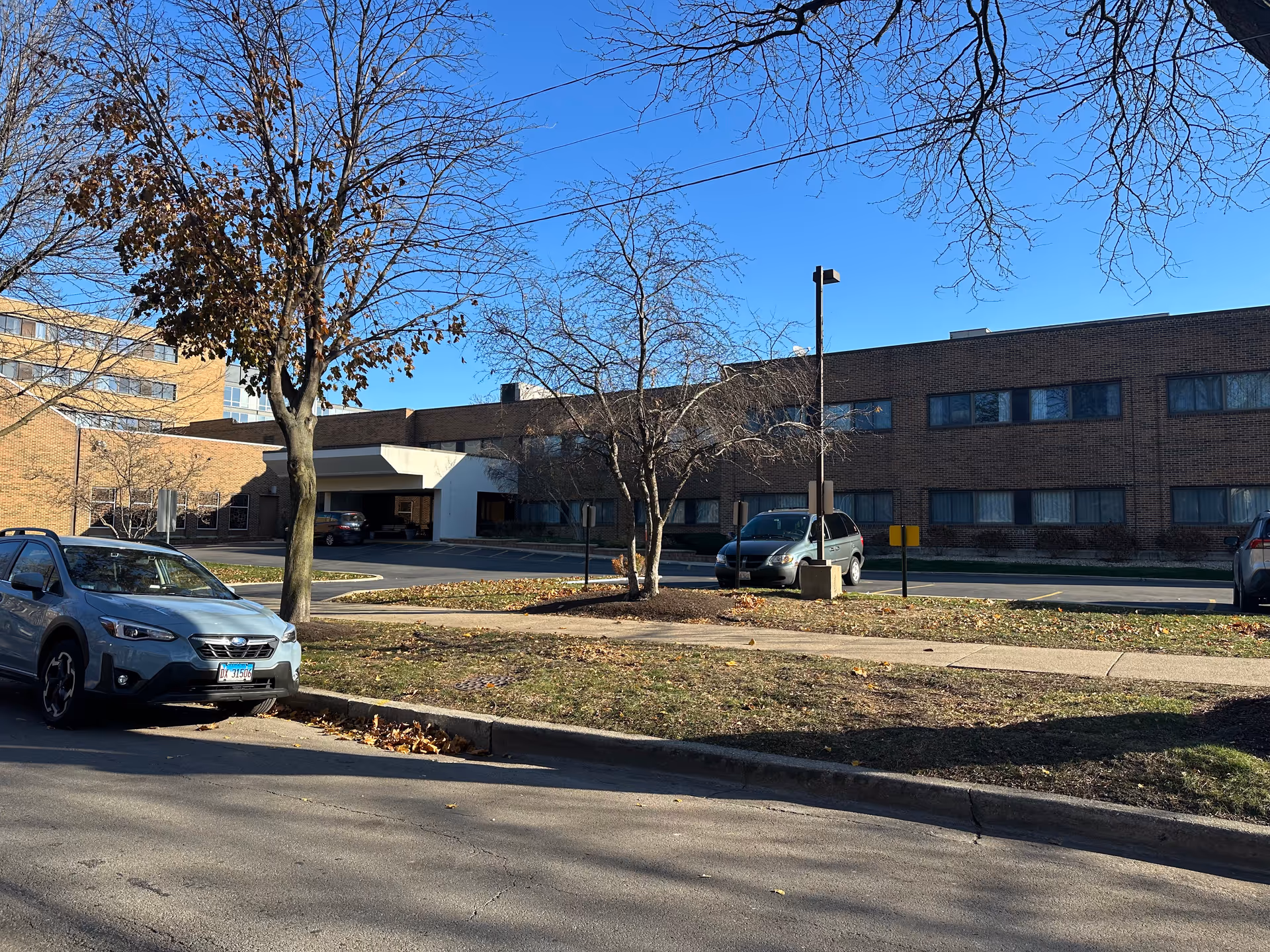 Exterior view of a two-story brick building with multiple windows, a covered entrance, and a parking area with several parked cars. Leafless trees and a clear blue sky are visible.