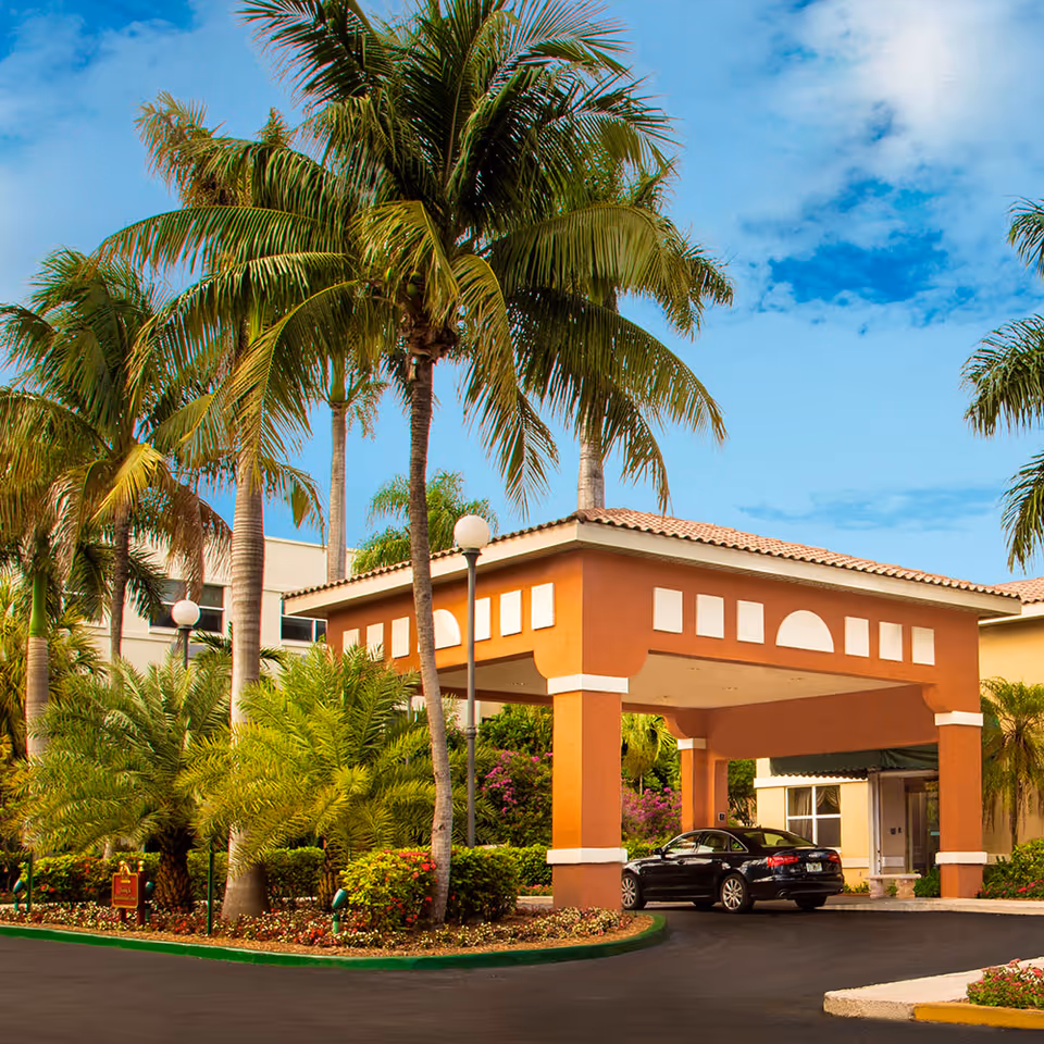 Entrance area of The Palace Renaissance & Royale facility with a covered driveway, surrounded by palm trees and lush landscaping under a partly cloudy blue sky. A black car is parked under the covered entrance.