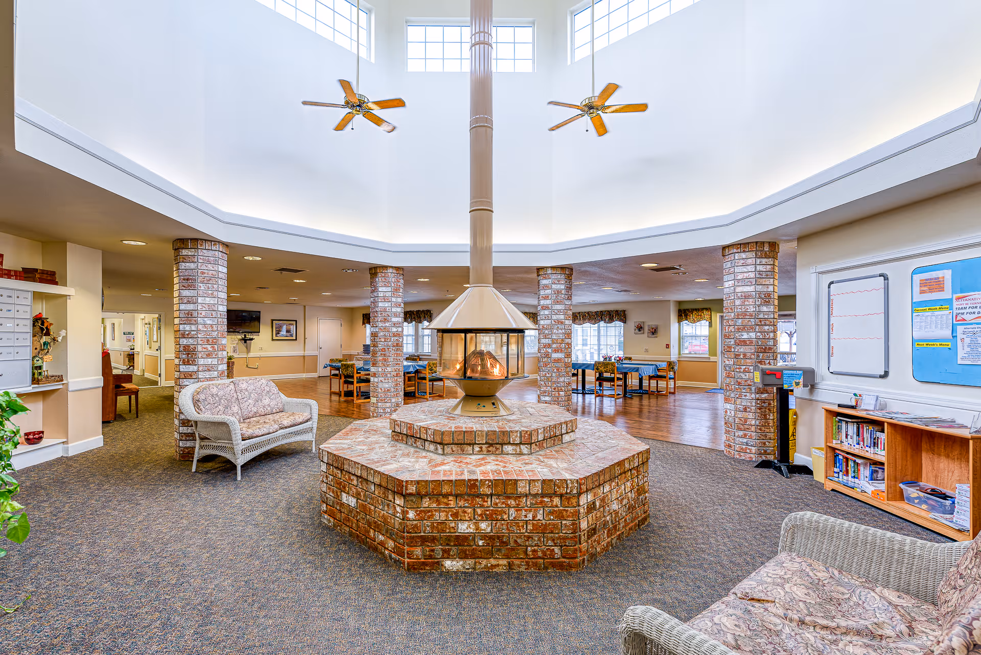 Spacious common area in an assisted living facility featuring a central brick fireplace with a tall chimney, surrounded by wicker seating with floral cushions. The room has high ceilings with large windows near the top, two ceiling fans, brick columns, and a carpeted floor. In the background, there are dining tables with chairs and windows letting in natural light.