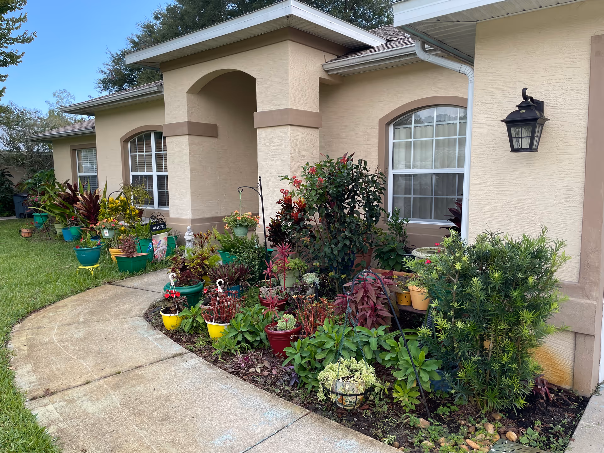 Exterior view of a beige single-story building with arched windows and a covered entrance. The front yard features a curved concrete walkway bordered by a garden bed filled with various green and flowering plants in pots and planted in the ground. A black outdoor lantern is mounted on the wall near one window.
