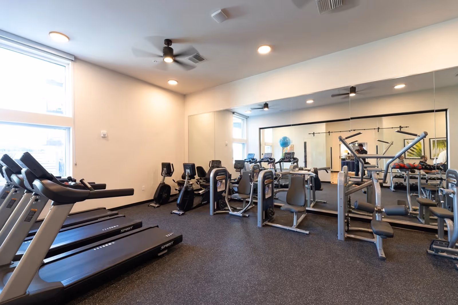 Well-lit fitness room with treadmills, exercise bikes, and strength machines reflected in a large mirror.
