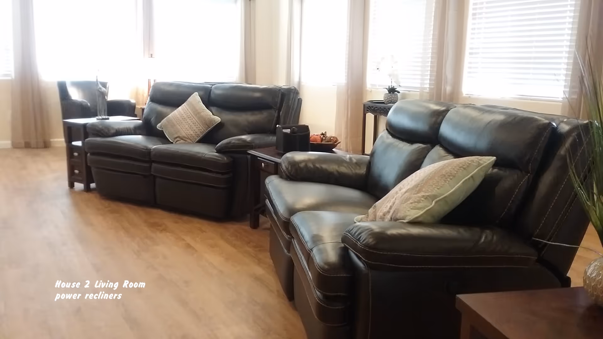 A living room with two black leather power recliner sofas, each with a decorative pillow. There are wooden side tables between and beside the sofas, one with a decorative bowl and another with a small plant. The room has large windows with white blinds and light-colored curtains, and a wooden floor.