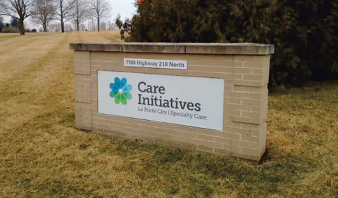 A brick sign on a grassy area with the text 'Care Initiatives La Porte City | Specialty Care' and an address plaque above it reading '1100 Highway 218 North'. Trees and open land are visible in the background.