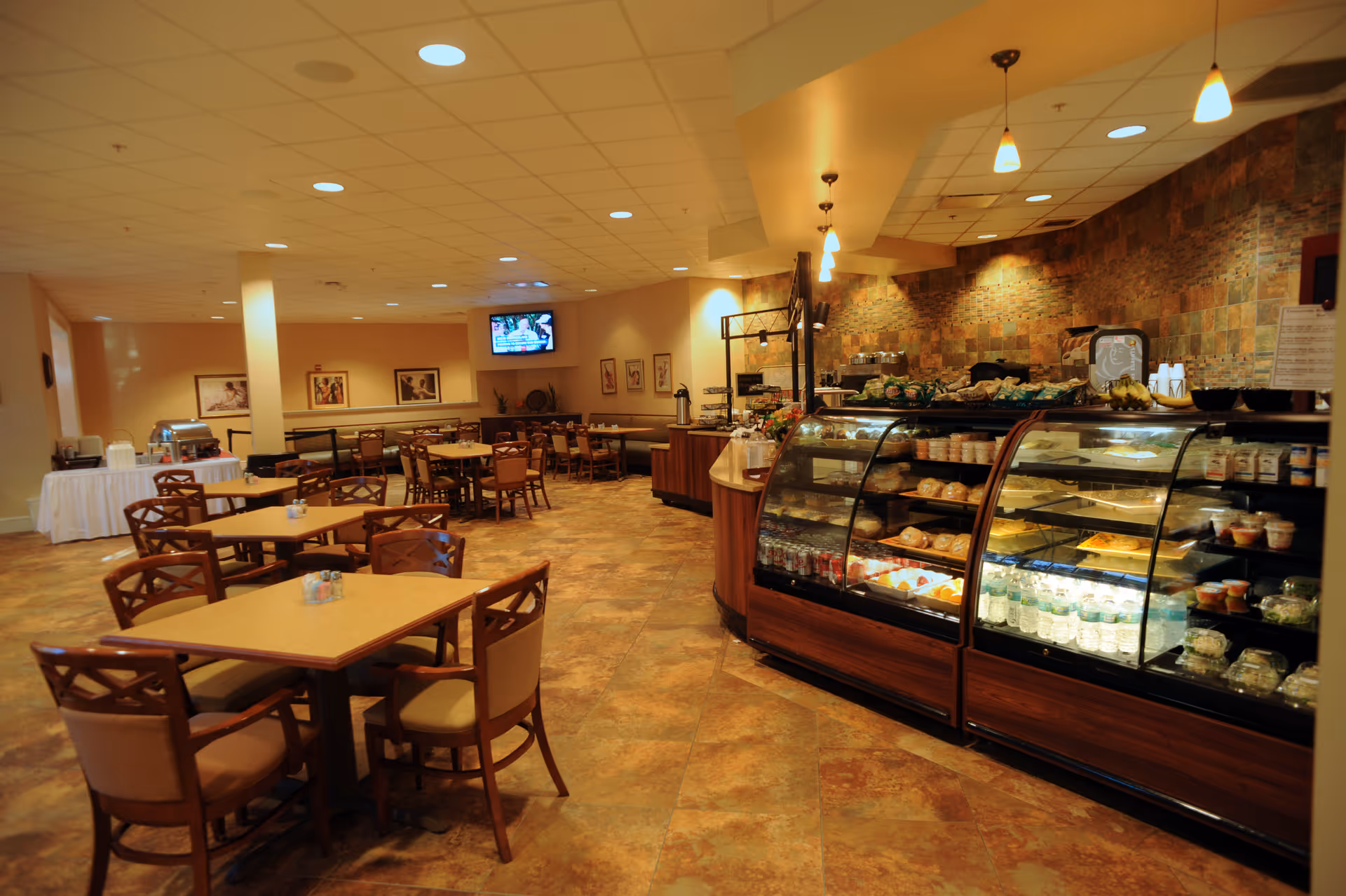 Dining room with multiple wooden tables and chairs and a refrigerated display case of food under warm pendant lighting.