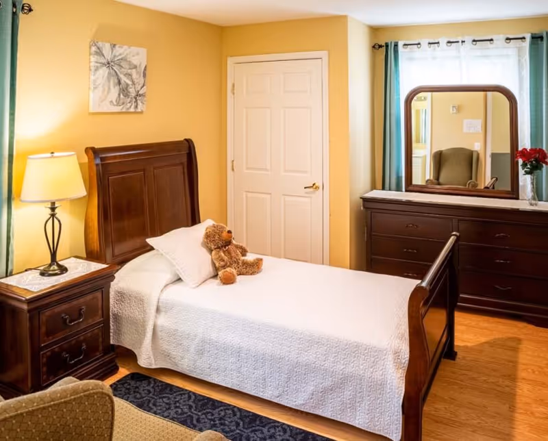 A cozy single bedroom with a wooden bed, nightstand and lamp, dresser with mirror, and a teddy bear on the bed.