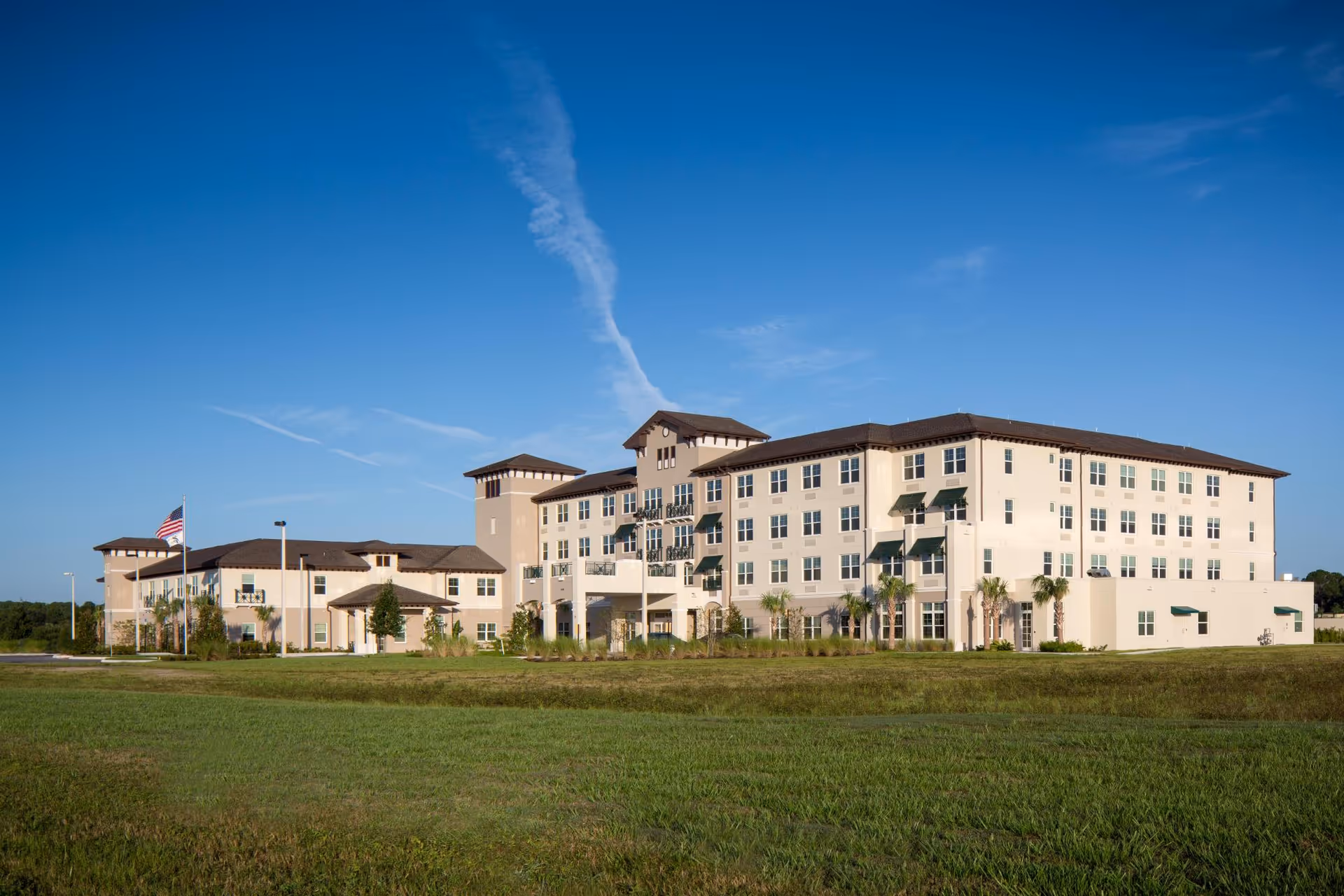Exterior view of a large, multi-story senior living facility building under a clear blue sky with an American flag on a flagpole in front and a well-maintained grassy lawn surrounding the building.