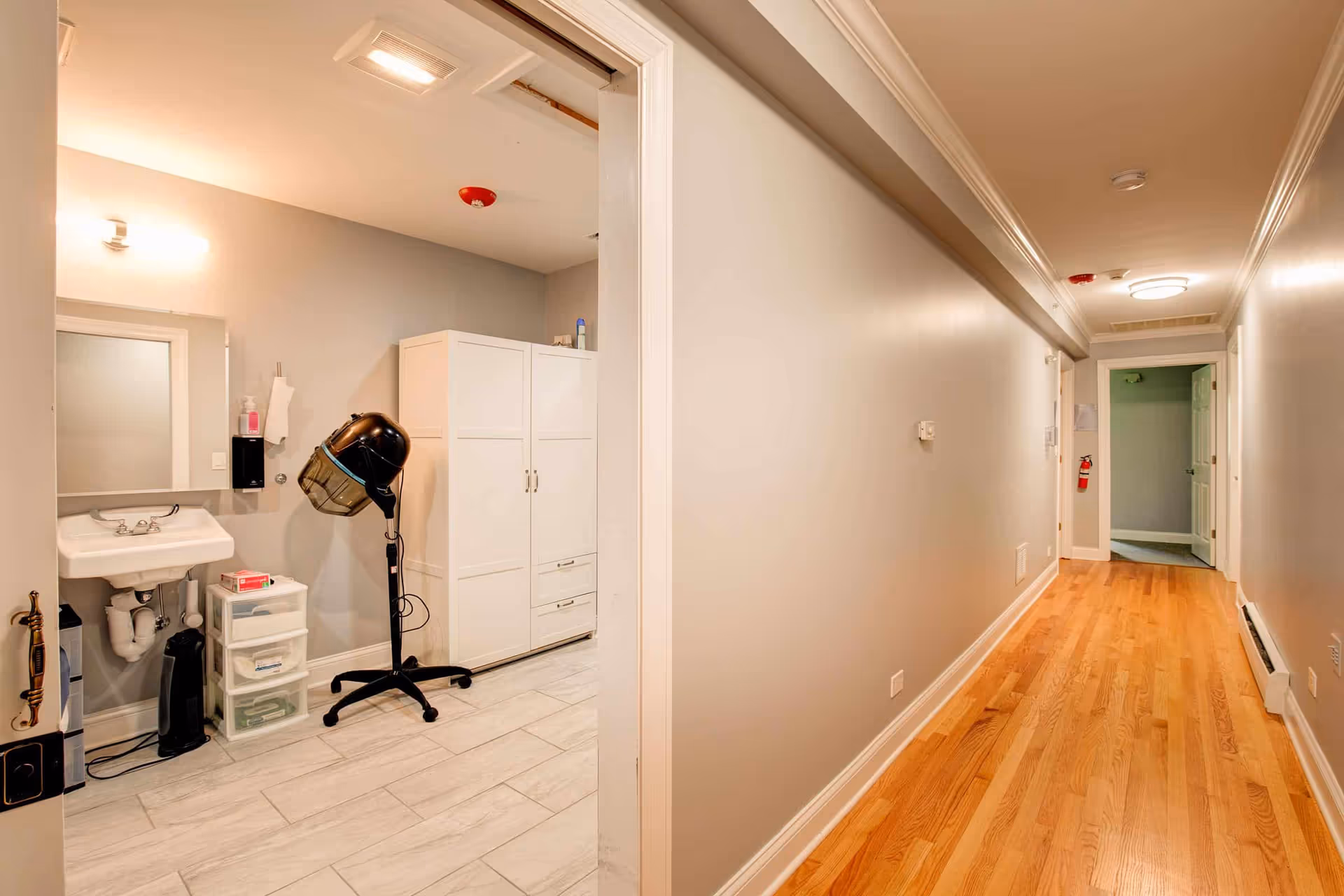 A hallway with wooden flooring and beige walls leading to an open door at the end. On the left side, there is an open room with tiled flooring containing a white sink, a mirror, a black hair dryer hood on a stand, a white storage cabinet, and a small plastic drawer unit.