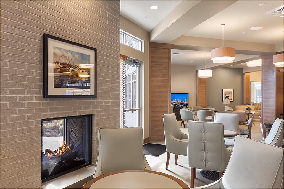 A cozy senior living facility common area with a lit fireplace set in a light brick wall, a framed painting of boats above it, and several round tables surrounded by cushioned chairs. The room has warm lighting with modern pendant lights and a TV visible in the background.