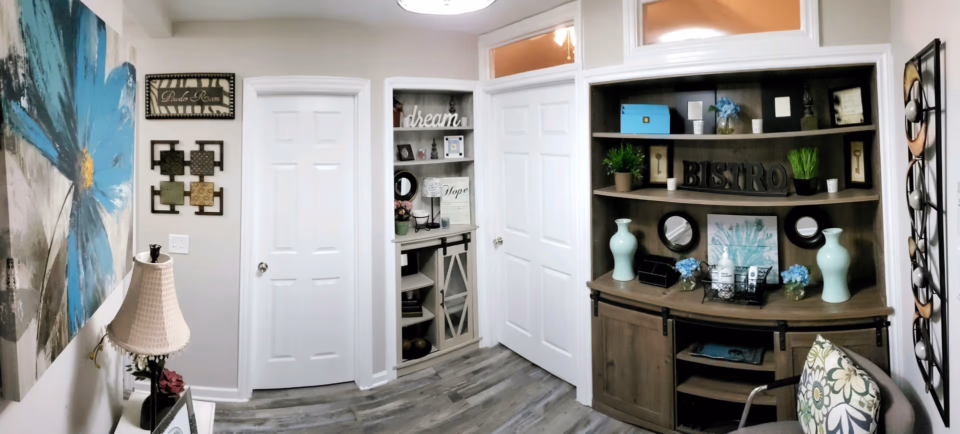 Interior view of a cozy senior living facility room with light-colored walls and wood flooring. The room features a large blue flower painting on the left wall, a table lamp with a beige shade, and decorative wall art. There are two white doors in the center, with a built-in shelving unit between them holding decorative items including a sign that says 'dream'. On the right side, a wooden cabinet with shelves displays various decor items such as vases, plants, and a 'BISTRO' sign. A chair with a floral cushion is partially visible in the foreground.