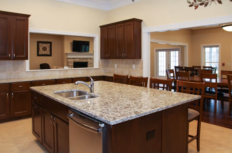 A spacious kitchen area with a large granite island countertop featuring a built-in sink and faucet. The kitchen has dark wooden cabinets and drawers, with a dishwasher integrated into the island. In the background, there is a dining area with multiple wooden chairs and tables, and a living room area with a fireplace and a wall-mounted TV visible through a large interior window.