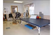 Physical therapy room with an exercise table, exercise balls, foam rollers, and various exercise equipment including a stationary bike and a chair. The room has a window with floral curtains and a mirror on the wall.