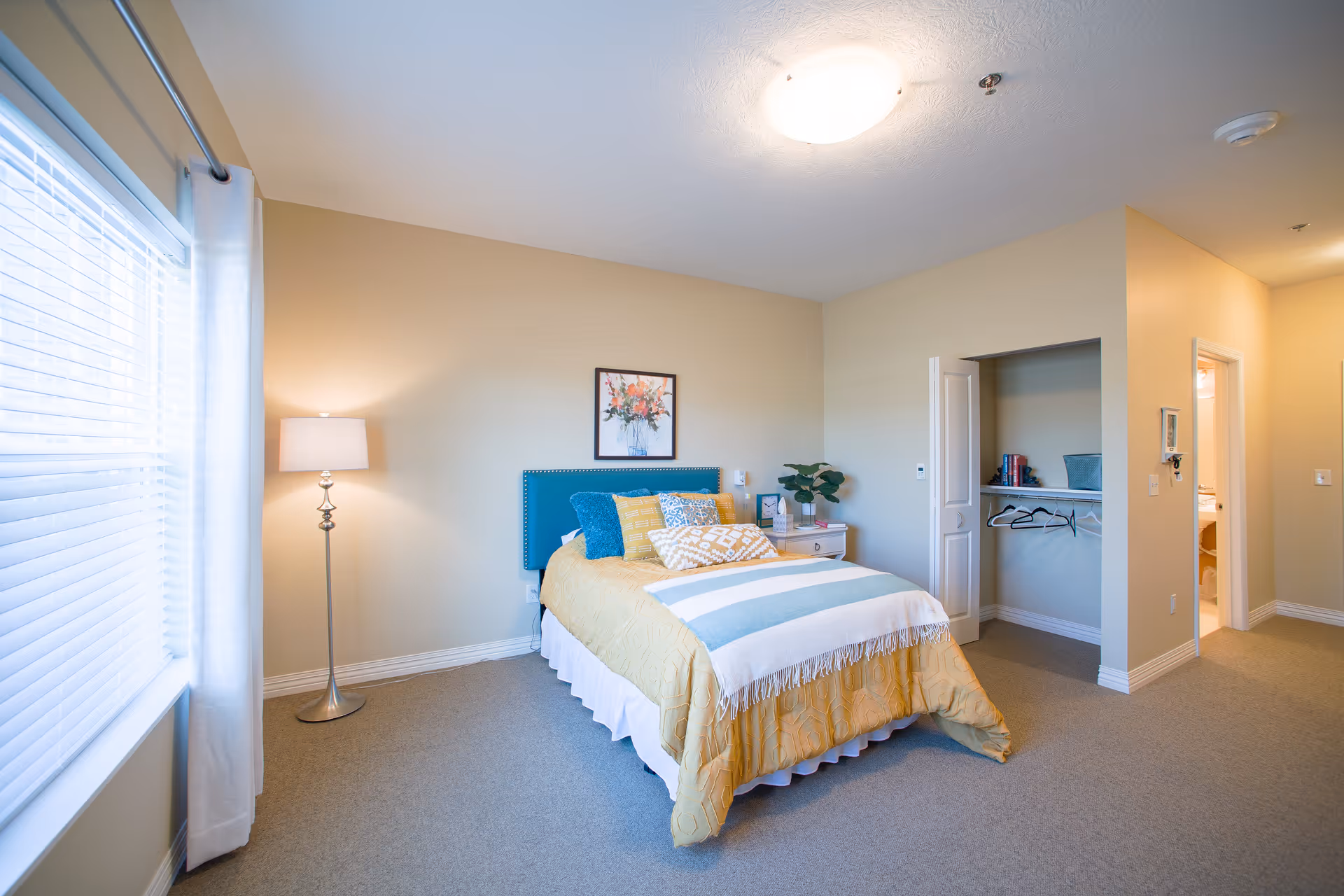 A bright and cozy bedroom with a bed featuring a teal headboard, yellow and blue bedding, and multiple pillows. There is a floor lamp next to a window with white blinds and curtains. A nightstand with a plant, clock, and books sits beside the bed. An open closet with hangers and shelves is visible, along with a doorway leading to a bathroom.