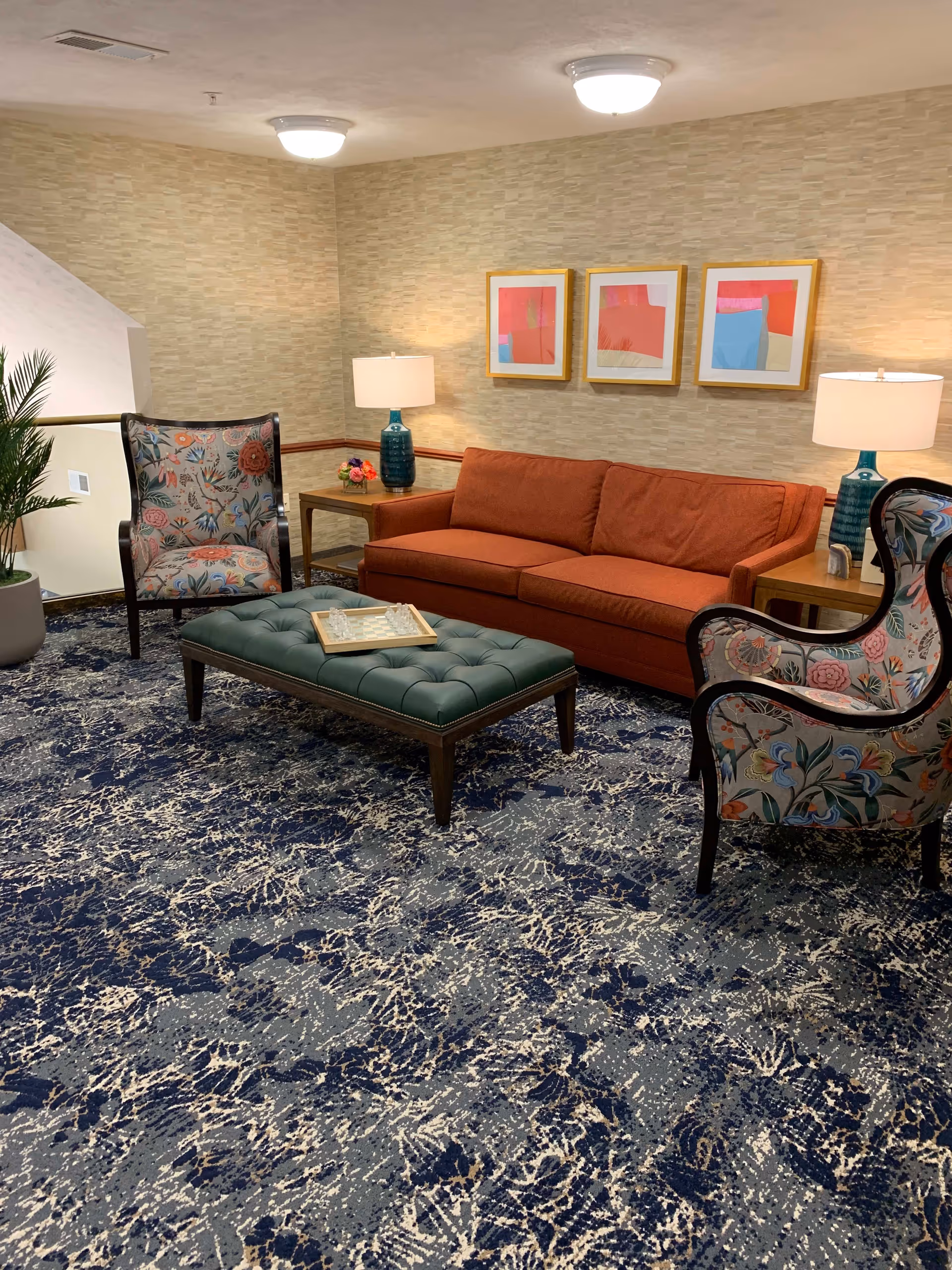 A cozy living room area with a burnt orange sofa, two floral-patterned armchairs, and a green tufted ottoman with a chessboard on top. The room has a blue and white patterned carpet, two side tables with teal lamps, and three colorful abstract paintings on the wall.