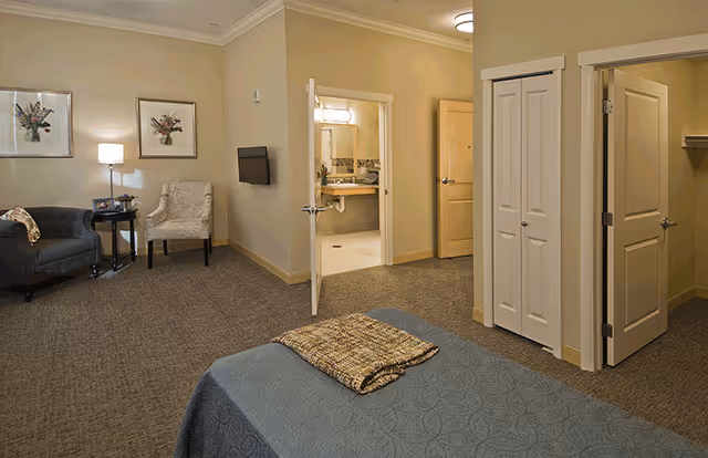 A furnished assisted-living bedroom with a bed in the foreground, a seating area to the left, and an open door revealing a bathroom and closets.