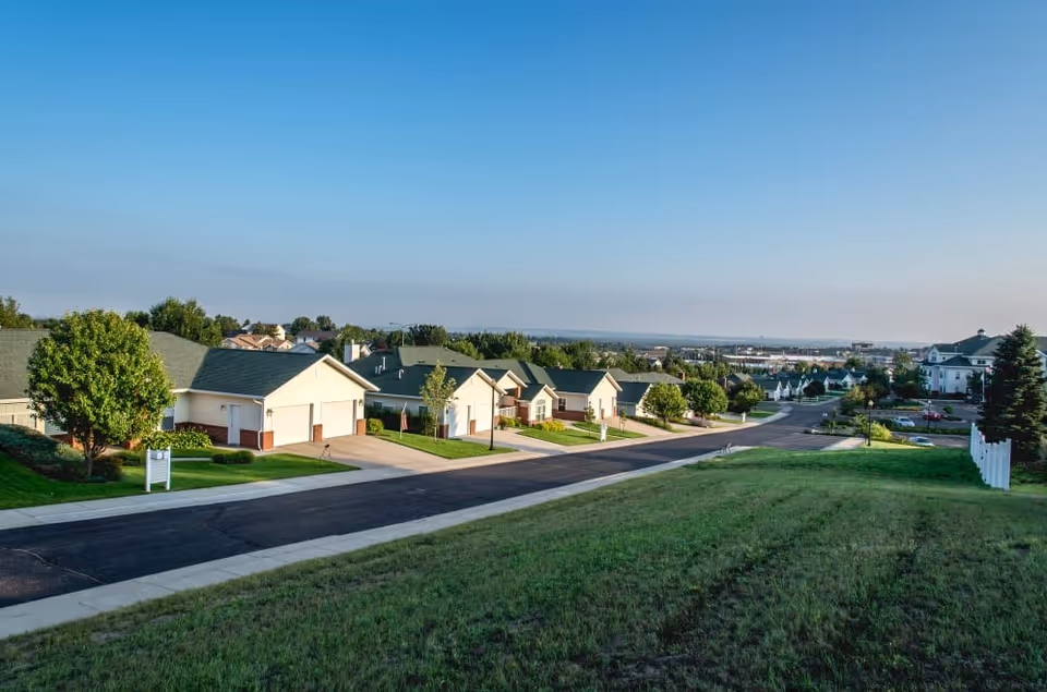 A scenic view of a residential neighborhood with single-story houses featuring green roofs and garages, a paved road running through the area, well-maintained lawns, trees, and a clear blue sky.