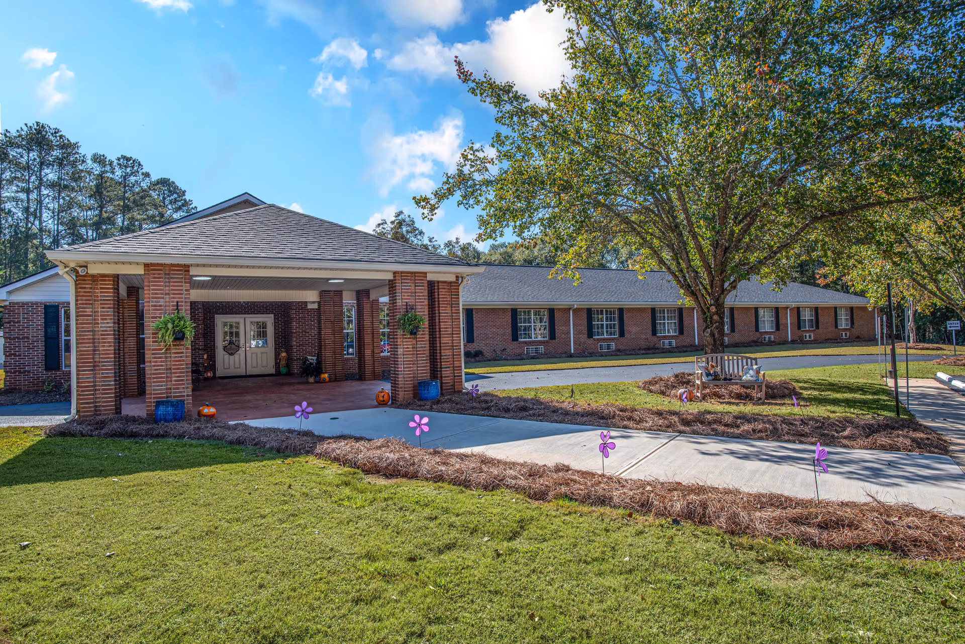 Front entrance of a single-story brick senior living facility with a covered entrance, landscaped walkway, and a large tree.