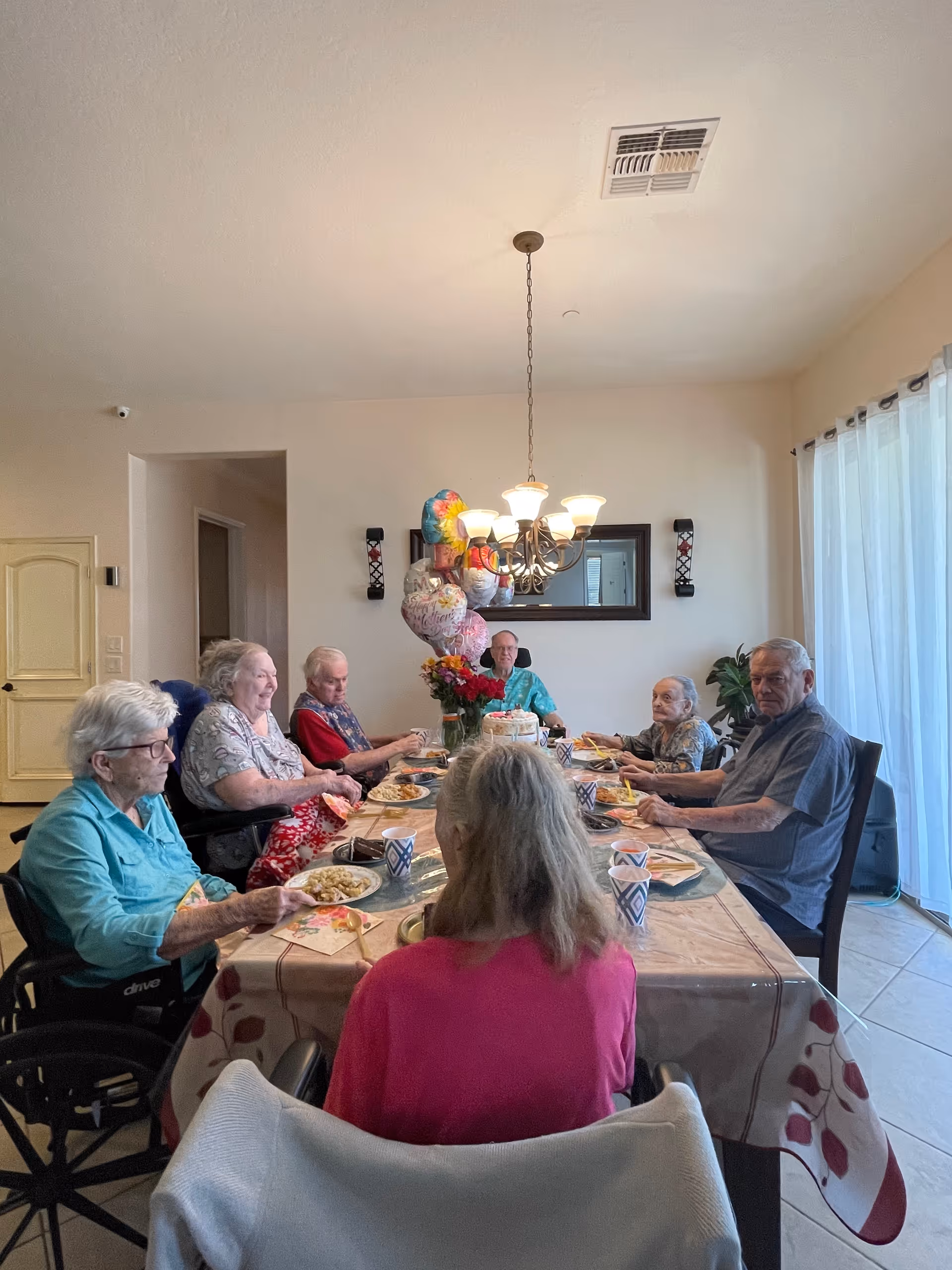 A group of elderly people sitting around a dining table in a well-lit room, enjoying a meal together. The table is covered with a floral tablecloth and has plates of food, cups, and a centerpiece with flowers and balloons. There is a chandelier hanging above the table and a large window with sheer curtains on the right side.