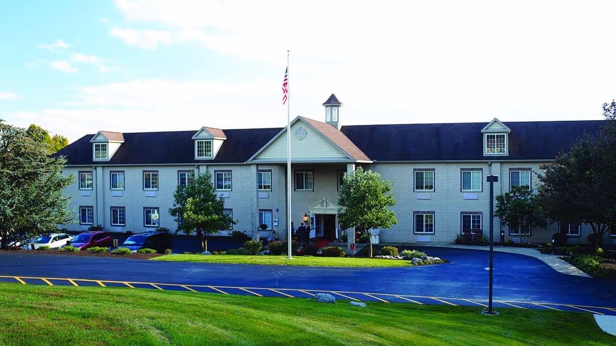 Exterior view of a two-story senior living facility building with a pitched roof, multiple windows, and a central entrance with a small portico. There is a flagpole with an American flag in front of the building, surrounded by green grass, trees, and a paved driveway with parked cars.