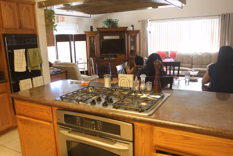 Open kitchen with a gas cooktop and oven facing a living area where several residents sit on sofas and chairs.