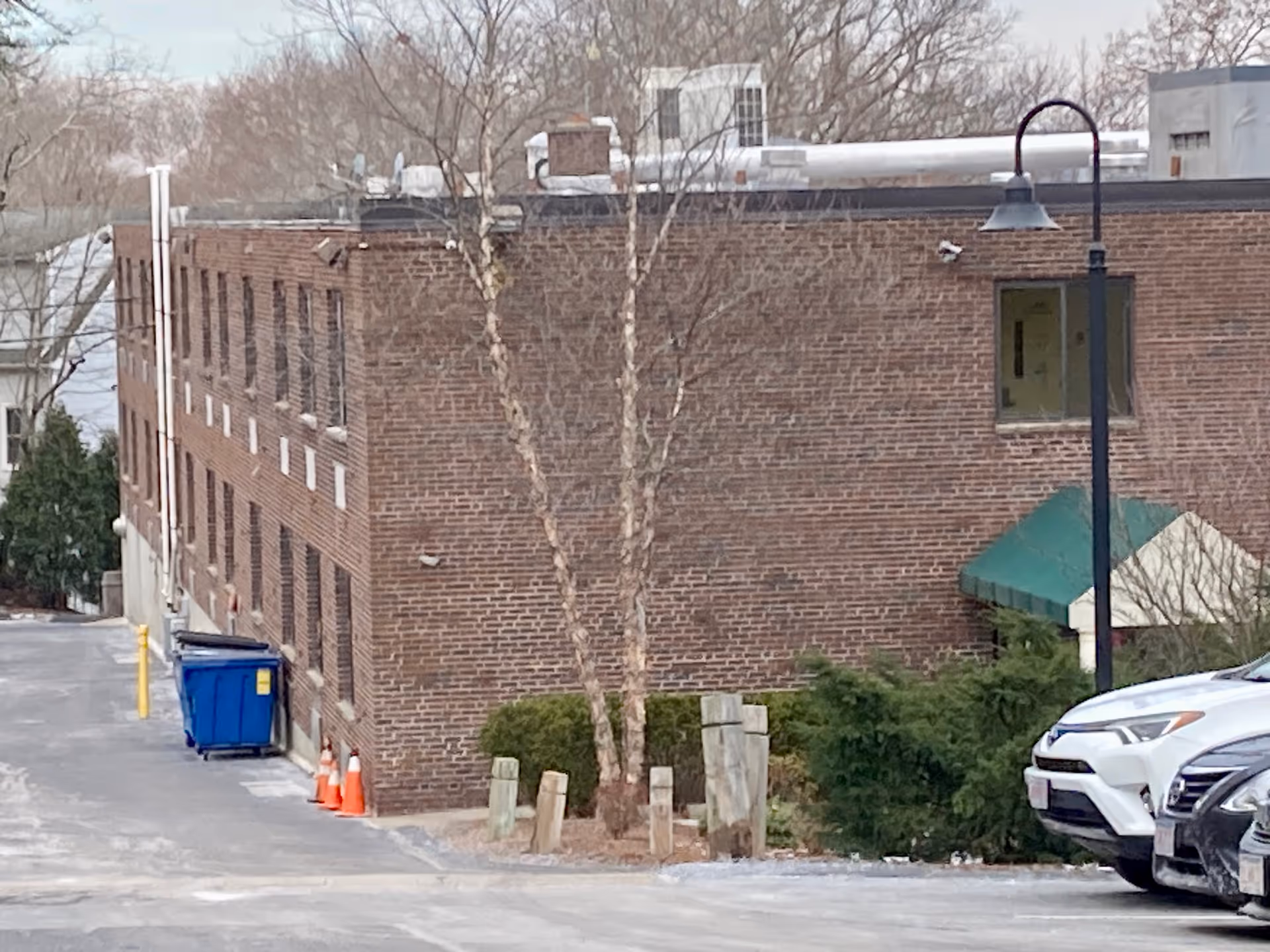 Exterior view of a brick building with a green awning over a door, a street lamp, several parked cars, some trees without leaves, orange traffic cones, and a blue dumpster along the side of the building.