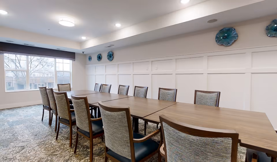 Long wooden dining table surrounded by chairs in a bright communal dining/meeting room with a large window and decorative wall plates.