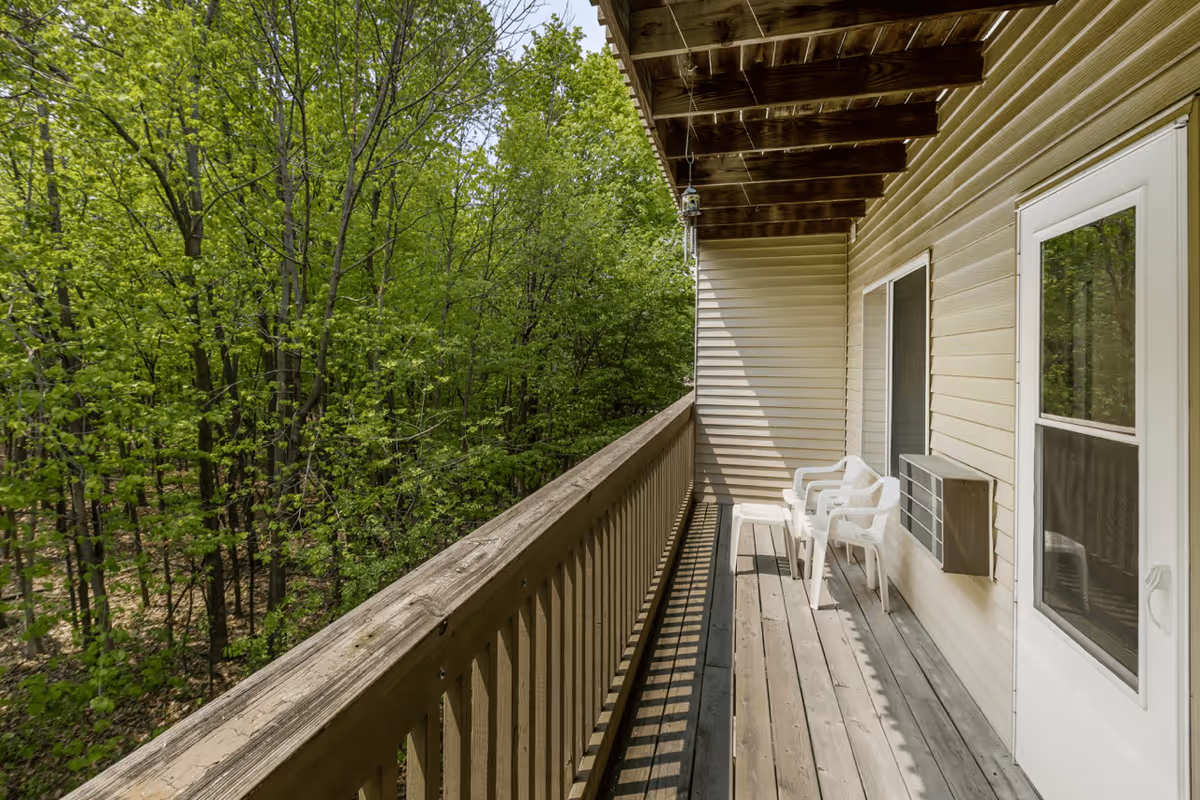 A wooden balcony attached to a beige siding building with two white plastic chairs and a small table. The balcony overlooks a dense green forest with many leafy trees. The balcony floor and railing are made of wood, and there is an air conditioning unit mounted on the wall next to a sliding glass door.