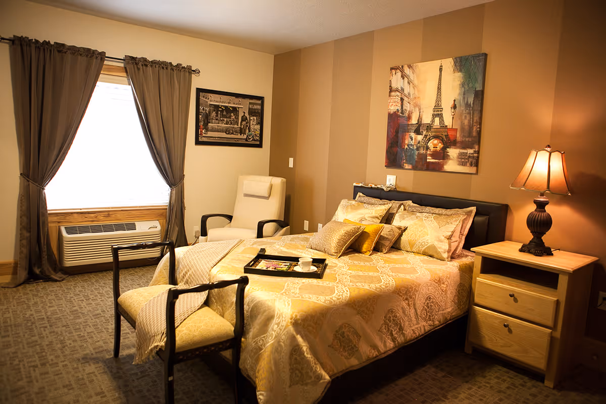A cozy bedroom with a neatly made bed featuring patterned gold and beige bedding. There is a wooden nightstand with a lamp on the right side of the bed. A cushioned chair is placed near the window, which is covered with brown curtains. The walls are painted in warm tones with a striped accent wall behind the bed, decorated with a framed picture of the Eiffel Tower. Another framed black and white photo hangs on the adjacent wall.