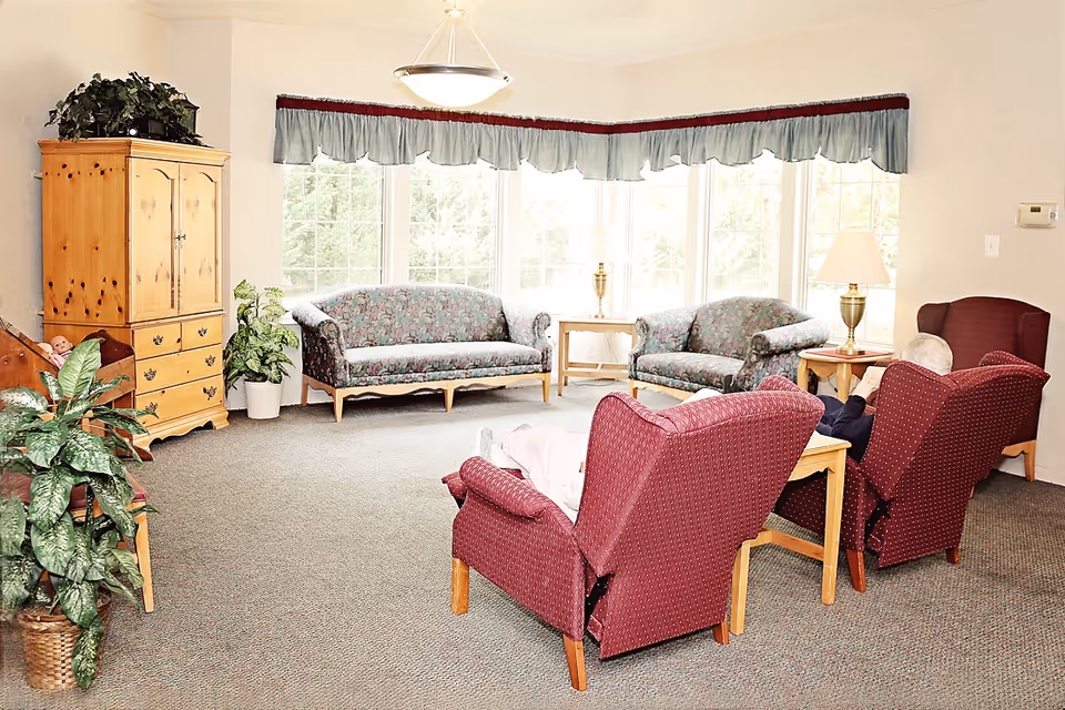 A bright living room in a senior care community with large windows letting in natural light. The room features two floral-patterned sofas against the windows, two red upholstered armchairs facing them, wooden side tables with lamps, a wooden cabinet with plants on top, and additional potted plants. Two elderly individuals are seated in the armchairs.