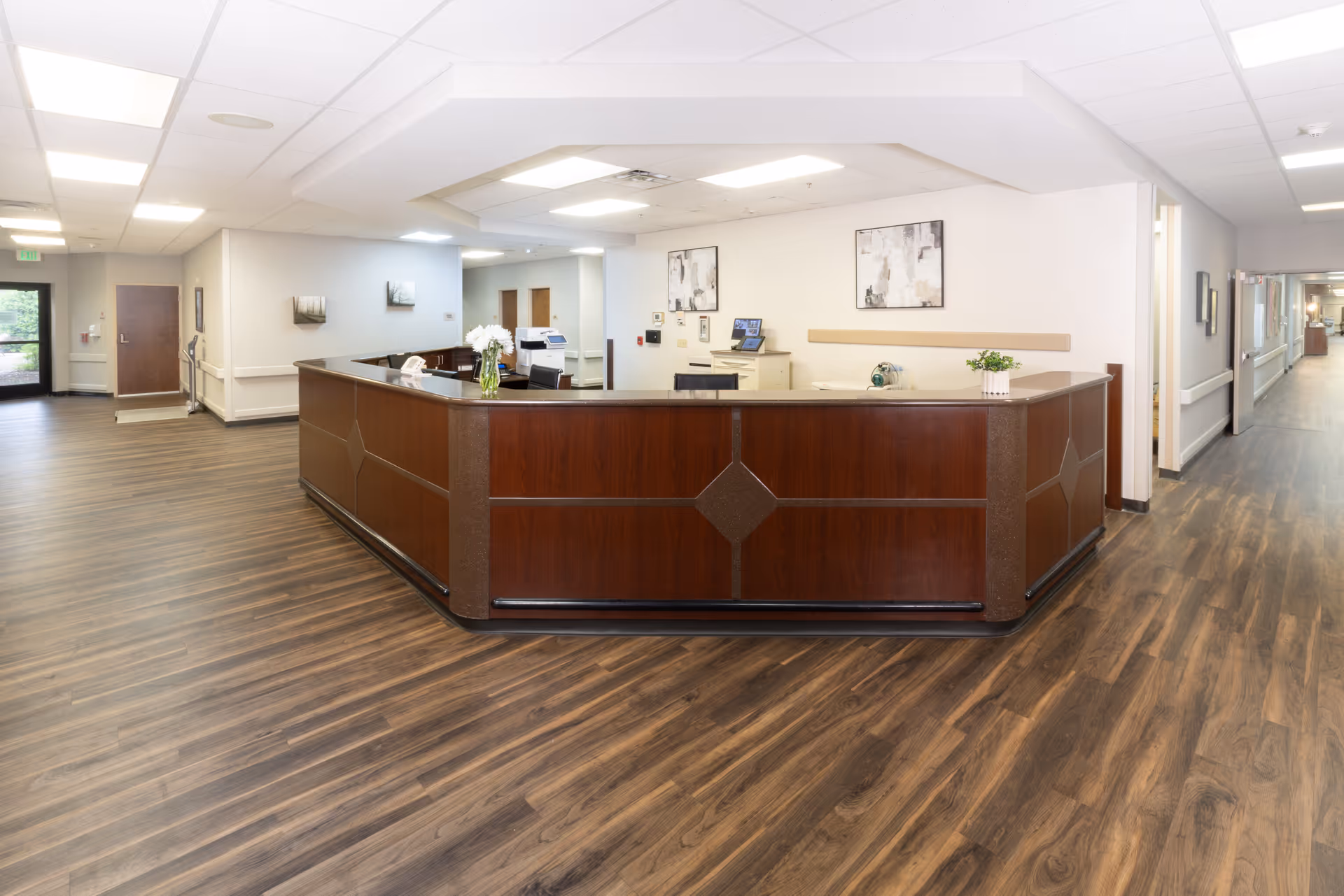 A spacious reception area in a senior living facility with a large wooden front desk in the center. The floor is covered with wood-patterned vinyl, and the walls are painted white. There are two abstract paintings on the wall behind the desk, a vase with white flowers on the desk, and a computer monitor. Hallways extend to the left and right, with doors and handrails visible along the walls.