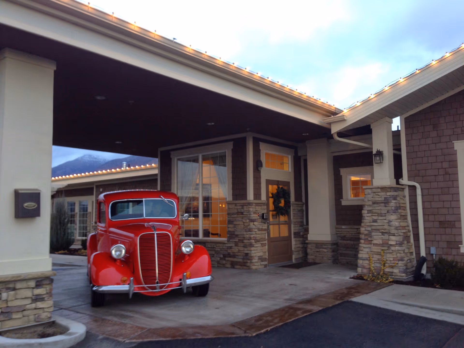 Exterior view of Ashford Assisted Living & Memory Care facility entrance with a covered driveway. A vintage red car is parked under the covered area near the front door, which has a wreath hanging on it. The building features stone and shingle siding with warm interior lights visible through the windows.