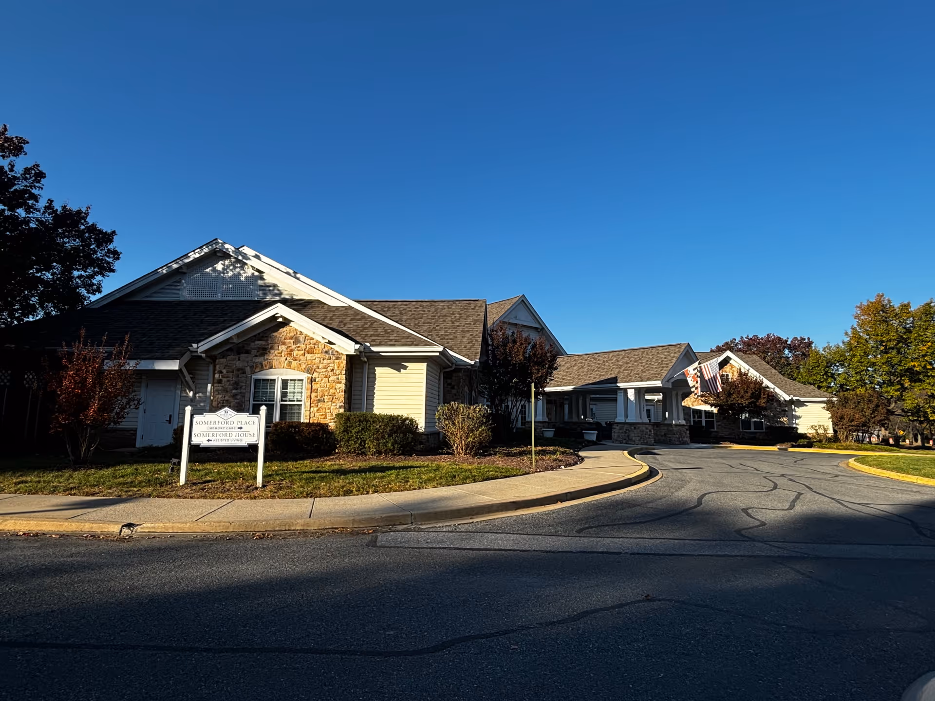 Exterior view of Somerford House Frederick, a single-story assisted living facility with a stone and siding facade, a covered entrance with an American flag, surrounded by trees and landscaping under a clear blue sky.