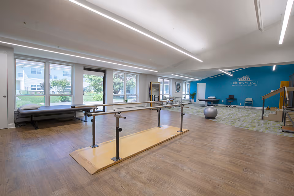 A spacious rehabilitation room with wooden flooring and large windows letting in natural light. The room features parallel bars for physical therapy, a therapy table with a pillow, an exercise ball, chairs, and a blue accent wall with the Friends Village Rehabilitation Services logo.