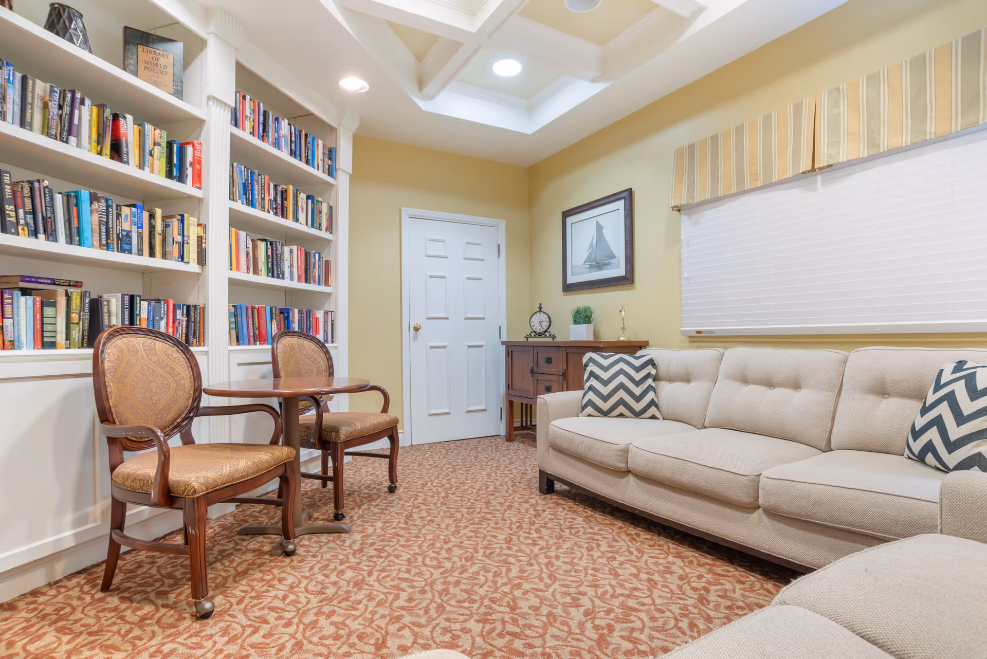 A cozy interior room with a beige couch adorned with chevron-patterned pillows, a wooden side table with a clock and plant, a framed picture of a sailboat on the wall, a window with closed blinds and a striped valance, and a built-in bookshelf filled with books next to two upholstered wooden chairs and a small round table. The room has a patterned carpet and a skylight in the ceiling.