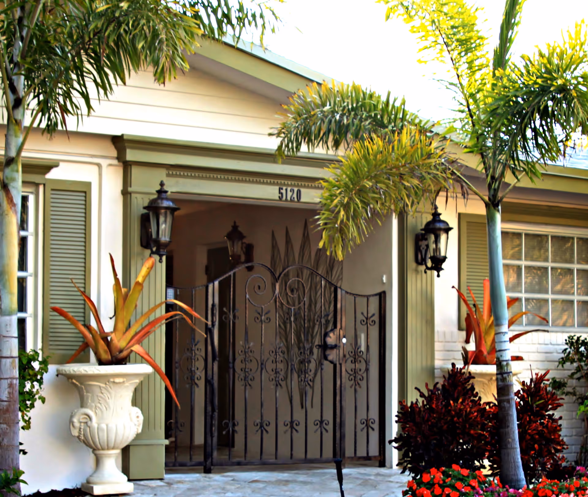 A decorative wrought-iron gate opens to a home's entrance, flanked by palm trees and potted plants.
