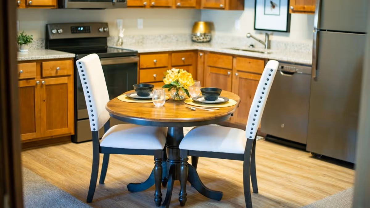 Round dining table set for two in an eat-in kitchen with wooden cabinets and stainless steel appliances.