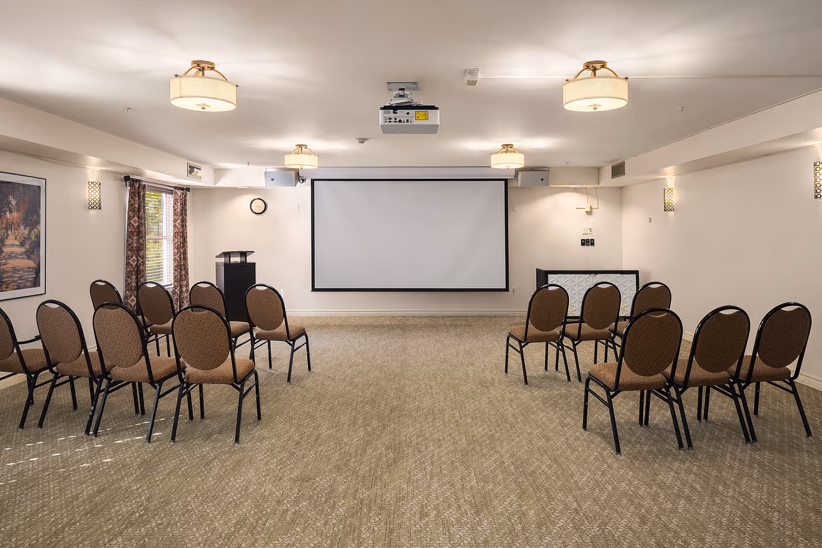 A conference or presentation room with rows of brown cushioned chairs arranged facing a large white projection screen on the wall. The room has beige carpeting, white walls, ceiling-mounted lights, a projector, a podium, and a window with patterned curtains on the left side.