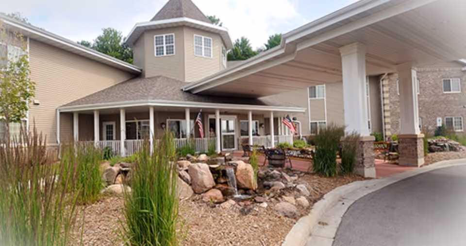 Exterior view of a senior living facility entrance with a covered driveway, landscaped garden featuring rocks and tall grasses, and two American flags near the entrance doors.