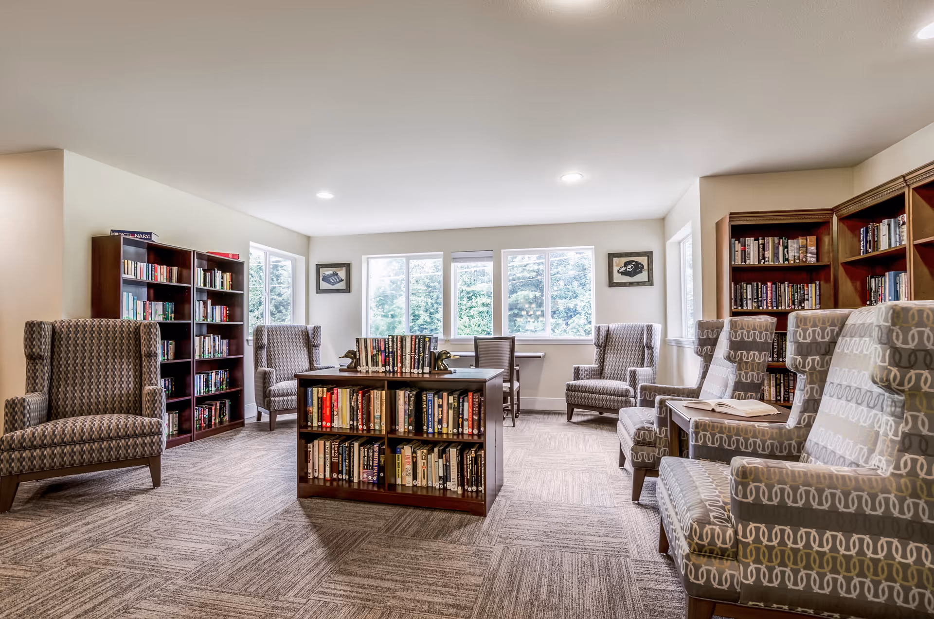 A bright and cozy library room with multiple bookshelves filled with books, several patterned armchairs arranged around the room, and a central bookshelf with books. Large windows let in natural light, and framed pictures hang on the walls.