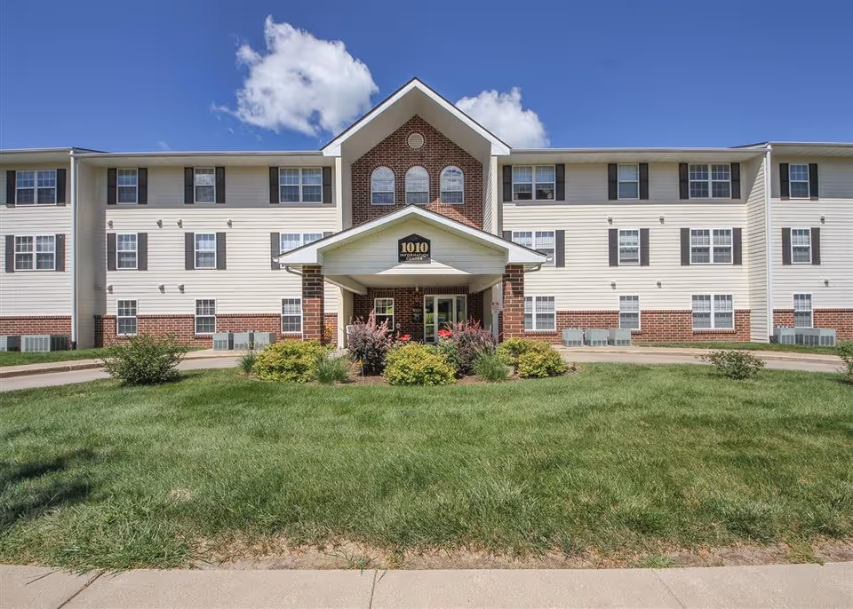Front exterior view of a three-story senior living facility building with a central entrance covered by a small gabled roof. The building has white siding with red brick accents and multiple windows. There is a well-maintained lawn and shrubs in front of the entrance under a clear blue sky with some clouds.