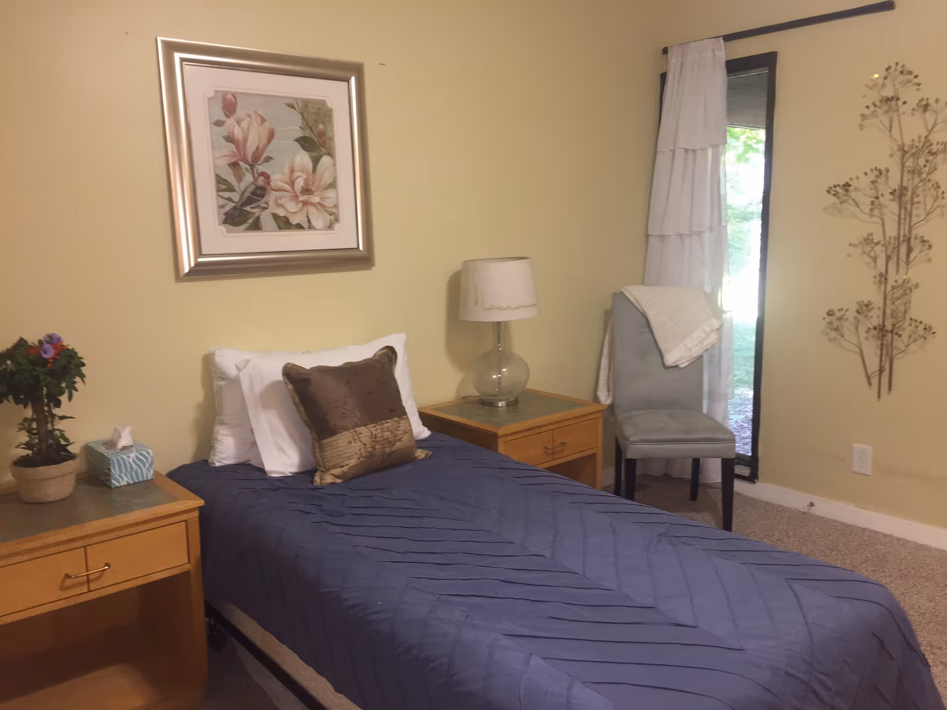 A small bedroom with a single bed covered with a blue quilt and two pillows, one white and one brown. There are two wooden nightstands on either side of the bed, one with a potted plant and a tissue box, and the other with a glass lamp. A framed floral and bird painting hangs on the wall above the bed. A gray upholstered chair with a white blanket draped over it is positioned near a tall window with white curtains. The walls are painted light yellow and there is a decorative metal wall art piece on the right side.