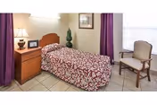 A small bedroom with a single bed covered in a red and white patterned bedspread. Next to the bed is a wooden nightstand with a lamp, a framed photo, and a small decorative plant. There is a wooden chair with a cushioned seat near a window with white blinds and purple curtains. The floor is tiled and the walls are light-colored.