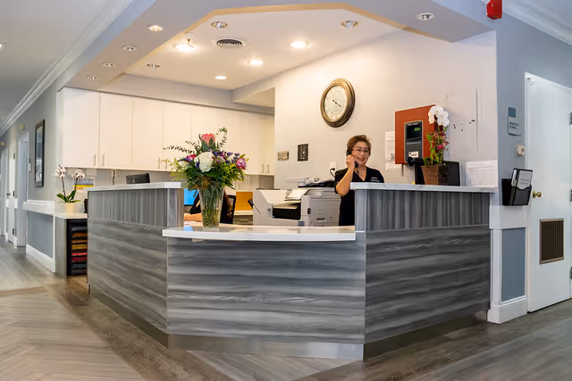 Reception area of a senior living facility with a woman standing behind a modern gray and white reception desk, talking on the phone. The desk has a vase with colorful flowers, and the background shows white cabinets, a clock on the wall, and office equipment.