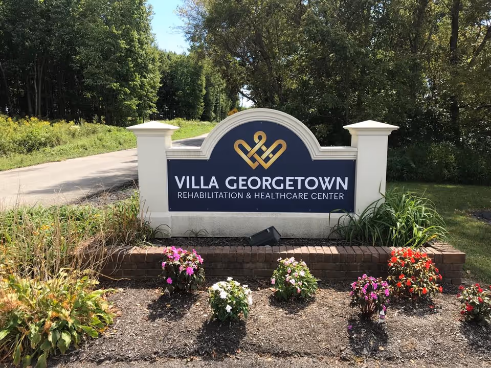 Outdoor sign for Villa Georgetown Rehabilitation and Healthcare Center surrounded by flowers and greenery, with a road and trees in the background.