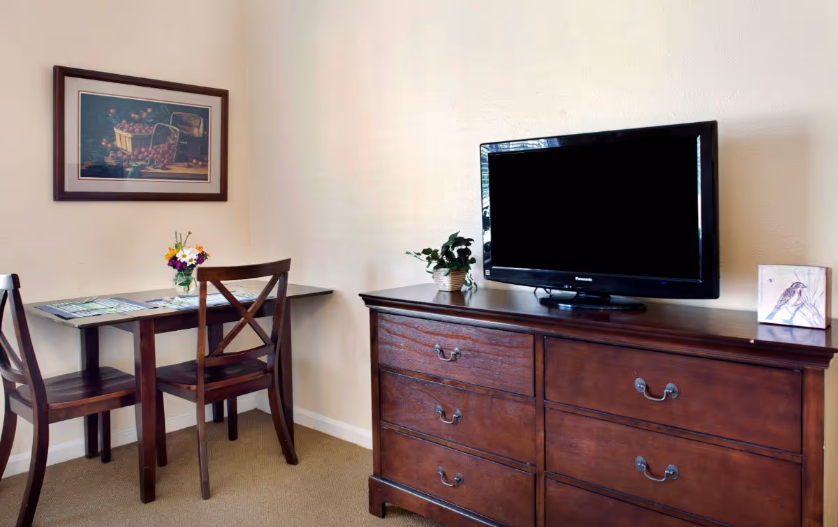 A small dining area with a dark wooden table and two matching chairs. On the table is a small vase with colorful flowers and some placemats. Above the table hangs a framed painting of baskets filled with fruit. To the right is a dark wooden dresser with a flat-screen TV on top, a small potted plant, and a decorative box with a bird illustration. The walls are light-colored and the floor is carpeted.
