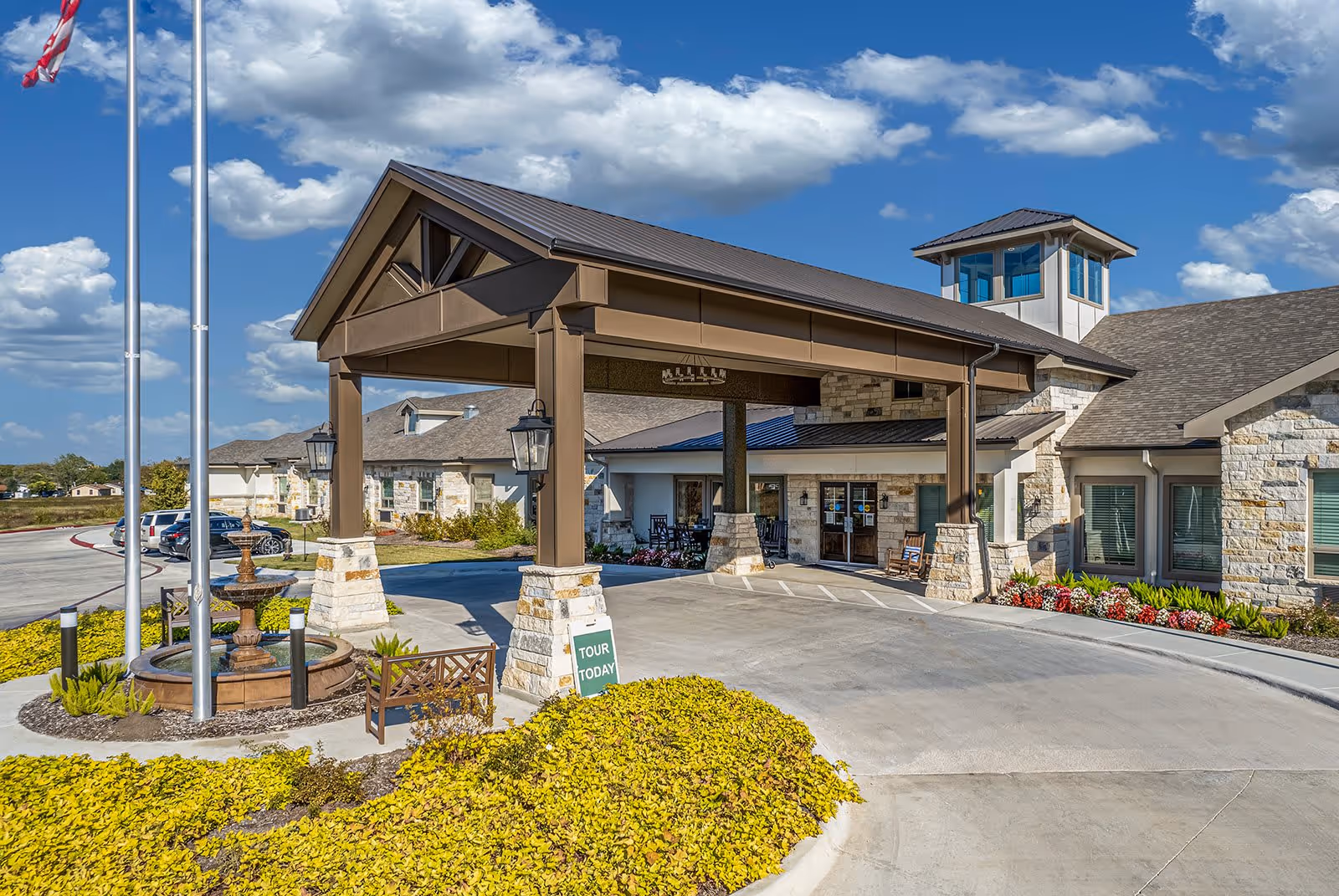 Entrance of Sage Valley Senior Living facility featuring a covered drop-off area with stone pillars and brown beams, a small fountain surrounded by yellow foliage, an American flag on a flagpole, and a clear blue sky with scattered clouds.