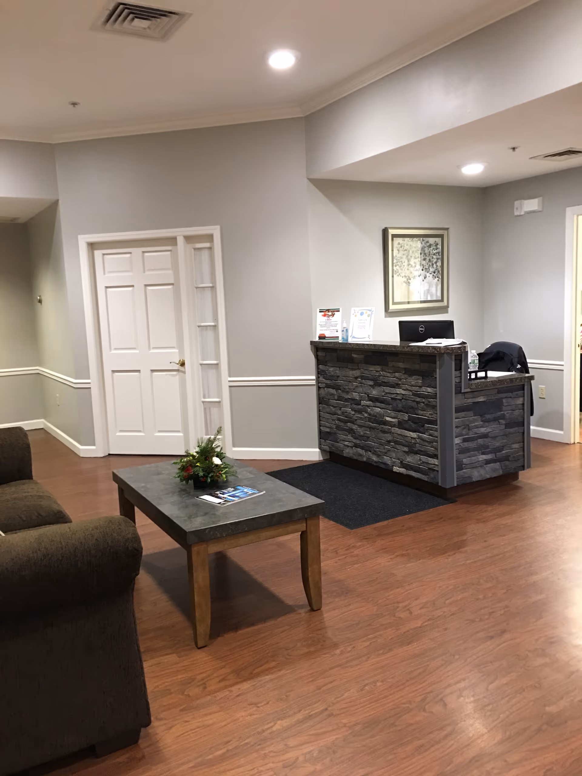 A neutral-toned reception lobby with a stone-front desk, coffee table with a small floral arrangement, and a couch on wood floors.